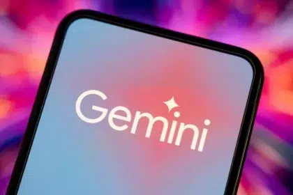 A smartphone screen displaying the "Gemini" logo in white text with a sparkle icon above the "i," set against a gradient blue and pink background. The phone is tilted, and the blurred background features vibrant shades of pink, purple, and orange.