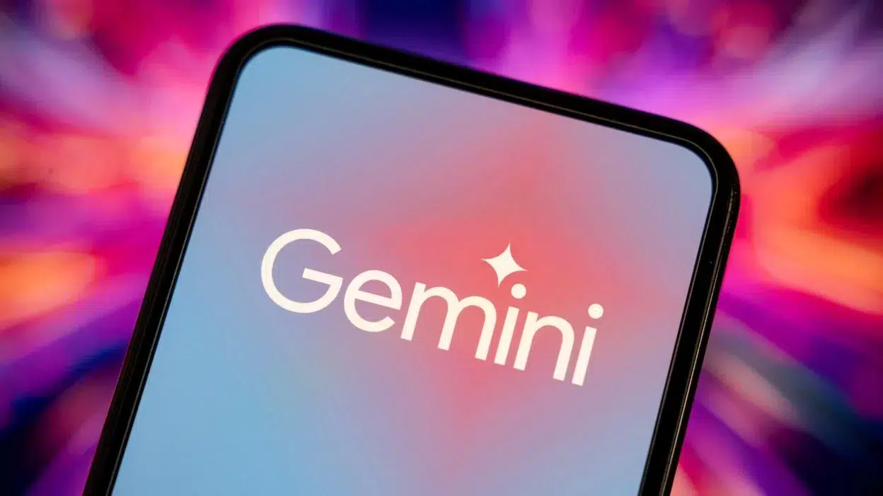 A smartphone screen displaying the "Gemini" logo in white text with a sparkle icon above the "i," set against a gradient blue and pink background. The phone is tilted, and the blurred background features vibrant shades of pink, purple, and orange.