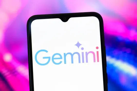 imageA smartphone displaying the "Gemini" logo in a blue-to-pink gradient with a sparkle icon above the "i," set against a vibrant, blurred pink and blue background.