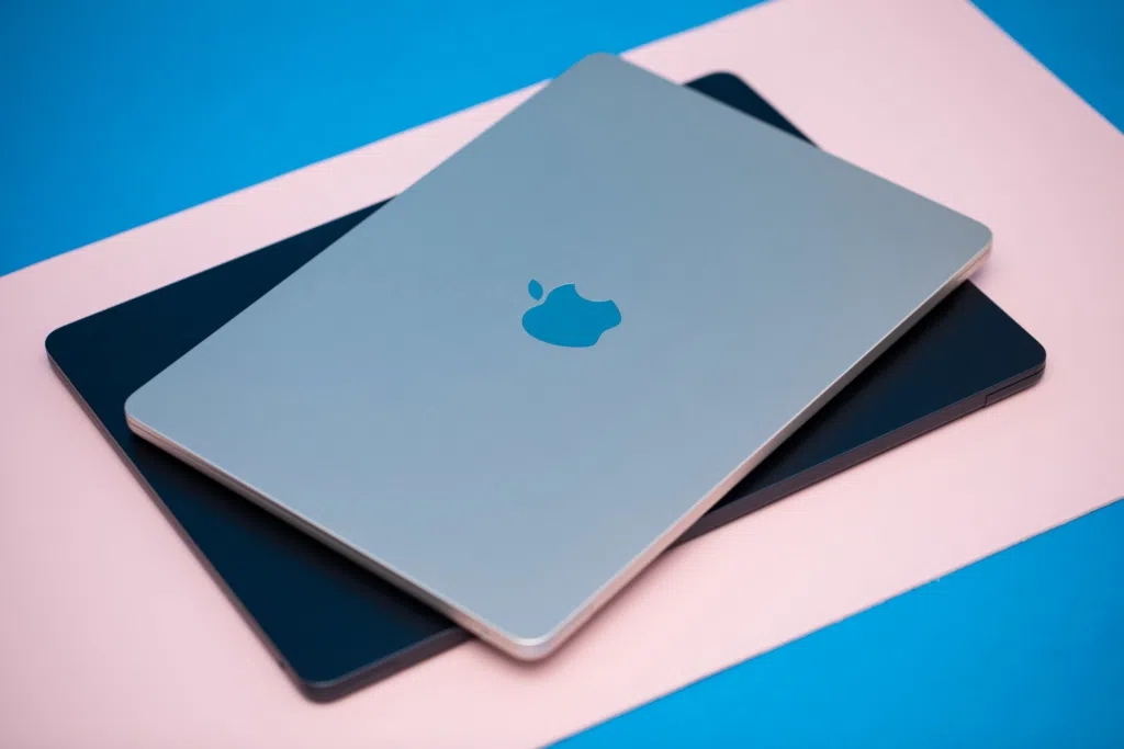 Two closed MacBook laptops, one in silver and the other in dark blue, stacked on top of each other on a pastel pink and blue background. The Apple logo is visible on the top device.